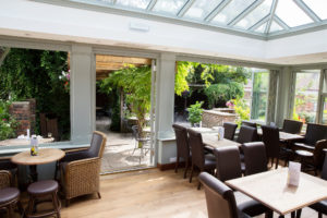 The Barn - Conservatory Bright, airy cafe with wooden tables, chairs, and lush greenery visible through large windows.